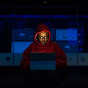 Focused woman in a red hoodie using a laptop in a dimly lit room with technology brands displayed.