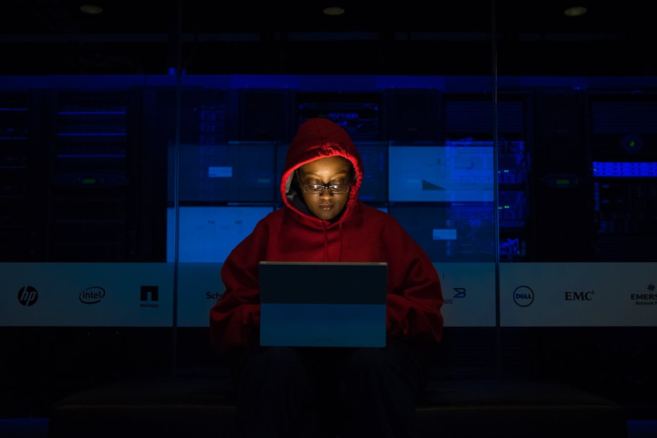services-01 Focused woman in a red hoodie using a laptop in a dimly lit room with technology brands displayed.