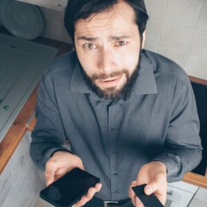 A confused man indoors looks up while holding a smartphone and credit card, concerned about a financial issue.