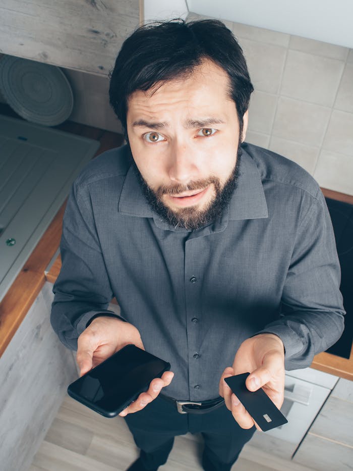 services-03 A confused man indoors looks up while holding a smartphone and credit card, concerned about a financial issue.